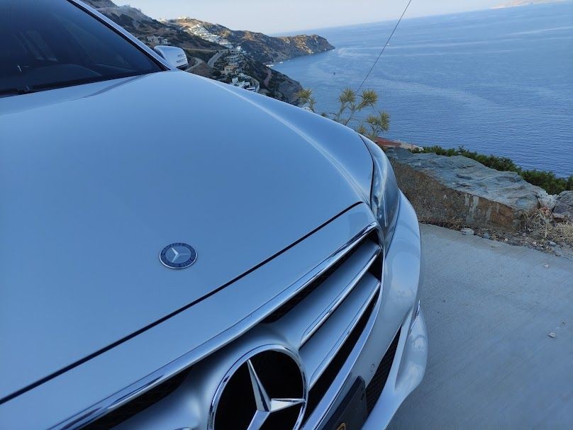 Close-up of a luxury car front grille and emblem with a scenic coastal view in the background.