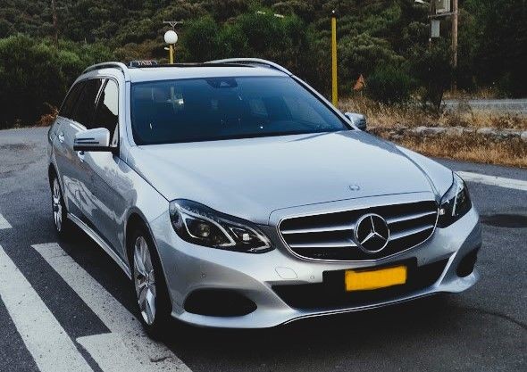 Silver Mercedes-Benz station wagon parked on a road with a scenic background of trees and hills.